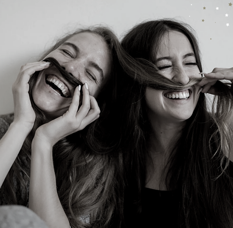 Das Bild ist schwarz-weiß und zeigt 2 Frauen, die gegenseitig ihre langen Haare nutzen, um sie sich wie Schnurrbärte auf die Lippe zu lagen. Sie lächeln breit und sehen aus als hätten sie jede Menge Spaß!