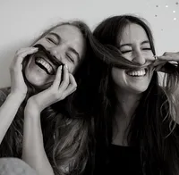 Das Bild ist schwarz-weiß und zeigt 2 Frauen, die gegenseitig ihre langen Haare nutzen, um sie sich wie Schnurrbärte auf die Lippe zu lagen. Sie lächeln breit und sehen aus als hätten sie jede Menge Spaß!