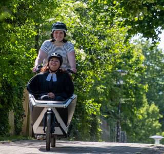 Eine Frau im gestreiften T-Shirt fährt auf einem Lastenrad einen Weg entlang. Im Hintergrund sind Bäume und Pflanzen. Vorne im Lastenrad sitzt eine andere Frau. Sie trägt einen Talar. Beide schauen fröhlich in die Kamera.