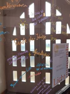 Eine Scheibe, auf die verschiedene Sätze und Wünsche zum Thema Licht geschrieben sind. Im Hintergrund eine 2flügelige Tür. Durch die vielen kleinen Fenster in der Tür dringt Licht und beleuchtet die Scheibe. Auf der Scheibe steht: Jesus ist das Licht der Dunkelheit,  Gerechtigkeit, meine Freunde sind mein Licht, und weiteres