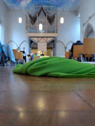 Im Vordergrund liegt ein grünes Tuch auf dem Boden einer Kirche, im Hintergrund sieht man verschwommen eine Orgel, vor der eine Person sitzt, ein Kreuz auf dem Altar, 2 Kerzen und 3 Lampen, rechts und links stehen Stühle und bilden einen Gang.