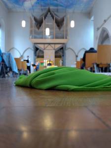 Im Vordergrund liegt ein grünes Tuch auf dem Boden einer Kirche, im Hintergrund sieht man verschwommen eine Orgel, vor der eine Person sitzt, ein Kreuz auf dem Altar, 2 Kerzen und 3 Lampen, rechts und links stehen Stühle und bilden einen Gang.